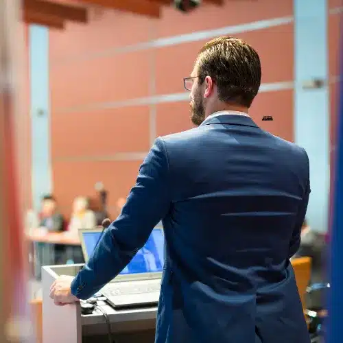 Man in a suit presenting at a cleaning business event, with audience in the background, emphasizing professional development and operational strategies for cleaning business owners.